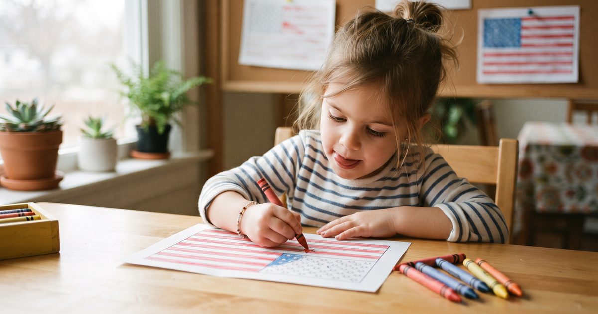 Close-up photo of a preschool-aged girl carefully coloring a small five-pointed star on a red, white, and blue flag coloring page with a red crayon, tongue poking out in concentration, a handful of sharpened crayons and a finished flag page nearby on a wooden table