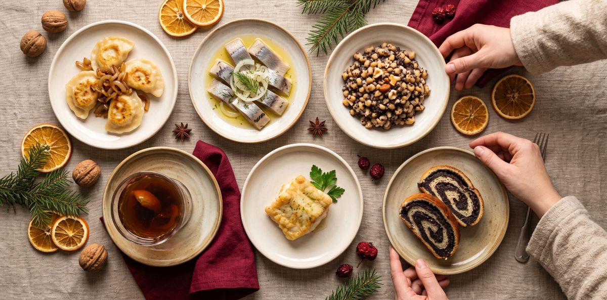 Overhead view of six small plates on a linen tablecloth: pierogi with fried onions, herring with onion, kutia, a glass of dried-fruit compote, baked fish and poppy seed roll, with dried orange slices, walnuts and fir branches